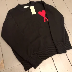 Svart stickad tröja från Ami Paris - Svart stickad tröja från Ami Paris med röd hjärtapplikation och bokstaven A på bröstet. Tröjan har rund hals, ribbade muddar och lång ärm. Perfekt för dig som gillar stilrena detaljer och vill ha något som sticker ut.