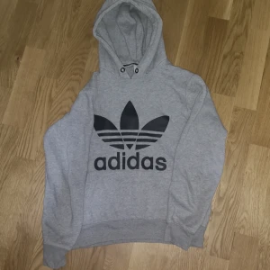 Grå Adidas hoodie med svart logga - Säljer en grå hoodie från Adidas med stor svart Trefoil-logga på bröstet. Tröjan har huva med snörning och är tillverkad i mjuk bomullsblandning. Perfekt för en chill och sportig stil. Långärmad och med ribbade muddar för extra komfort.
