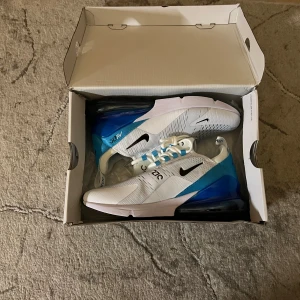 Nike Air Max 270 vit/blå sneakers - Nike Air Max 270 sneakers i vit och blå med svarta detaljer. Snygg mesh-överdel, synlig Air-enhet i hälen och snörning med blå accenter. Platt sula och rund tå ger en sportig vibe. Perfekt för dig som gillar streetstyle och komfort. Storlek 42