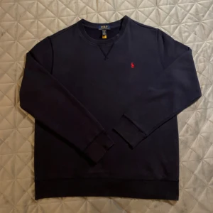Mörkblå sweatshirt från Polo Ralph Lauren - Mörkblå sweatshirt från Polo Ralph Lauren i storlek XL (I barnstorlek så motsvarar XS-S i vanlig storlek). Klassisk crewneck med röd broderad logga på bröstet. Tröjan har långa ärmar, ribbade muddar och är tillverkad i mjuk bomull. Använd fåtal gånger och väldigt fint skick.