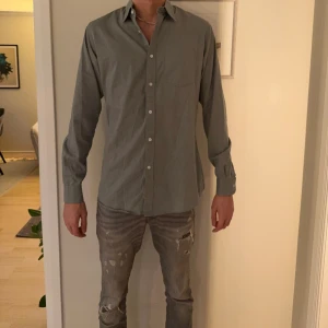 Grågrön skjorta med knappar - Snygg grågrön skjorta med klassisk krage och knappar hela vägen framtill. Skjortan har lång ärm med manschett och en diskret bröstficka. Passformen är normal och materialet känns som bomull, perfekt för en clean och stilren look.