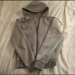 Grå hoodie från Polo Ralph Lauren - Snygg grå hoodie från Polo Ralph Lauren med broderad logga på bröstet. Tröjan har huva med snörning, dragkedja framtill och ribbade muddar vid ärmslut och nederkant. Perfekt för chill dagar och enkel att matcha med jeans eller joggers.