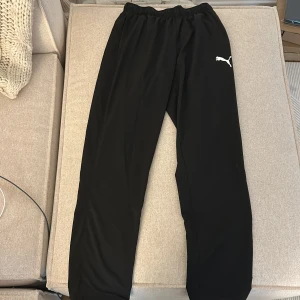 Svarta träningsbyxor från Puma - Svarta träningsbyxor från Puma med elastisk midja och klassisk vit Puma-logga på låret. Byxorna är tillverkade i ett lätt och smidigt syntetmaterial som passar perfekt för träning eller chill. Raka ben och enkel design för en clean look.