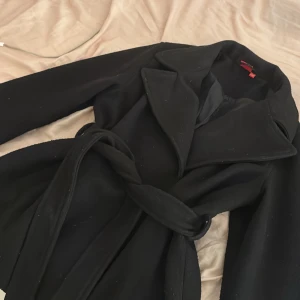 Svart kappa med bälte från RYVLS - Säljer en svart kappa från RYVLS med bred krage och knytbälte i midjan. Jackan har lång ärm och en stilren, klassisk look som passar perfekt till jeans eller dressade byxor. Materialet känns mjukt och har en snygg struktur.Kappan är lite större än storlek 146/152, skulle säga att den motsvarar ungefär storlek XS eller S.