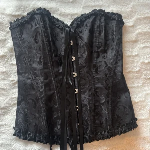 Svart korsett med snörning och volang - Säljer en svart korsett i storlek S med snyggt mönstrad yta och glansig finish. Korsetten har snörning både fram och bak, dekorativa volanger längs över- och nederkant samt metallhakar framtill. Materialet är polyester och elastan för en tight passform. Kontakta mig för fler bilder💞 perfekt inför halloween💞