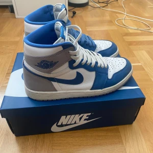 Nike Air Jordan 1 High True Blue - Säljer ett par Nike Air Jordan 1 High i blått, vitt och grått skinn. Klassisk siluett med högt skaft, perforerad tå och snörning. Ikonisk vinge-logga på sidan och Nike Air-märke på plösen. Kommer med originalkartong och extra skosnören. Pris går att diskutera.