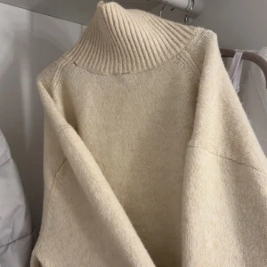 Beige polotröja i ull - Mysig beige polotröja med ribbad hög krage och långa ärmar. Tröjan är stickad i mjuk ull och har en loose passform som ger en chill vibe. Perfekt för kalla dagar när du vill vara både bekväm och snygg.