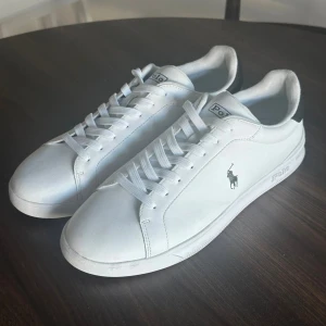 Vita Ralph Lauren sneakers - Snygga vita sneakers från Polo Ralph Lauren med klassisk design. Skorna har vit ovandel i skinn, svart detalj på hälen och Polo-logga på sidan. Platt sula och snörning framtill. Perfekta för en clean och stilren look.