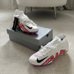 Nike Zoom Superfly 9 Elite FG fotbollsskor Storlek 41 - Nike Zoom Superfly 9 Elite FG fotbollsskor i vitt med svarta och röda detaljer. Skorna har snörning, Dynamic Fit och en cool grafisk design på sidorna. Yttersulan har lila och röda inslag med Zoom-teknologi för extra grepp på planen. Tillverkade i syntetmaterial för lätt känsla.