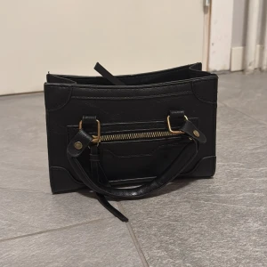 Svart handväska med gulddetaljer - Stilren svart handväska i skinnimitation med rektangulär form och snygga guldfärgade metalldetaljer. Väskan har praktiskt handtag och en dragkedja framtill för extra förvaring. Perfekt storlek för det viktigaste och ger en cool vibe till din outfit.