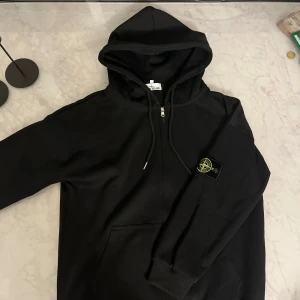 Svart hoodie från Stone Island - Svart hoodie från med klassisk logotyp-patch på vänster ärm. Tröjan har dragkedja framtill, huva med snörning och ribbade muddar. Tillverkad i mjuk bomull, perfekt för chill dagar. Snygg och enkel design som alltid funkar.