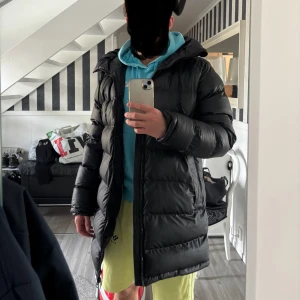 Svart pufferjacka från Everest - Säljer en svart pufferjacka från Everest med huva och dragkedja framtill. Jackan har breda, quiltade paneler, två stora fickor och justerbar dragsko i huvan. Perfekt för kalla dagar när du vill hålla värmen och ändå se stilren ut. Jätte bra skick knappast använd. Köpte den för 1999 