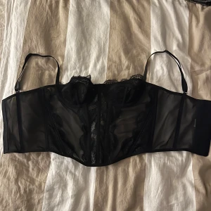 Svart spetskorsett med meshdetaljer - Säljer en svart korsett med genomskinliga meshpaneler och vacker spets längs kuporna och framsidan. Toppen har justerbara axelband och hakknäppning bak för perfekt passform. Snygg v-ringning och detaljerad spets ger en lyxig vibe.