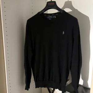 Stickad Polo Ralph Lauren tröja - Svart långärmad tröja från Polo Ralph Lauren i slim fit med broderad logga på bröstet. Liten i storlek så nassar både M/L.Tillverkad i mjuk pima bomull som känns skön mot huden. Klassisk rund hals och ribbade muddar vid ärmslut och nederkant. Perfekt för dig som gillar stilrena och enkla plagg.