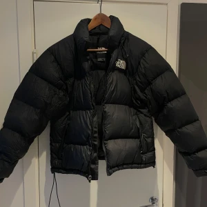 Svart pufferjacka från The North Face - Säljer en svart pufferjacka från The North Face i storlek S. Jackan har hög krage, synlig logga på bröstet och dragkedja framtill. Den är vadderad och har en loose passform med långa ärmar. Perfekt för kalla dagar och har en clean, urban stil.