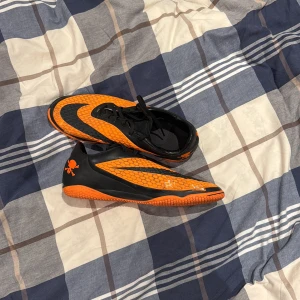 Nike fotbollsskor svart/orange  - Säljer ett par Nike fotbollsskor i svart och orange med coolt bikakemönster och dödskalle-detalj på hälen. Skorna har snörning och platt sula, perfekta för inomhusspel. Materialet är syntet och designen är riktigt sportig och edgy.