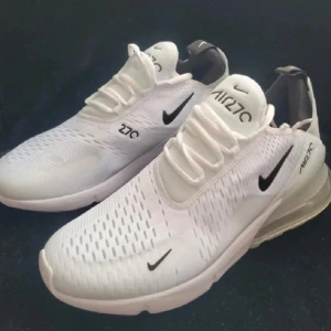 Nike Air Max 270 vita sneakers - Nike Air Max 270 sneakers i vitt med svarta detaljer och stor synlig Air-enhet i hälen. Ovandelen är i mesh för bra andningsförmåga och har snörning framtill. Snygg och sportig design med rund tå och platt sula, perfekt för dig som gillar streetwear.