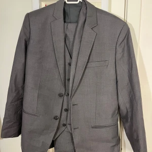 Grå kostym från Ram Fashion - Snygg grå kostym från Ram Fashion med klassisk kavaj och matchande väst. Kavajen har två knappar, smala slag och flera fickor. Västen har fyra knappar och enkel design. Perfekt för dig som vill ha en stilren och tidlös look.