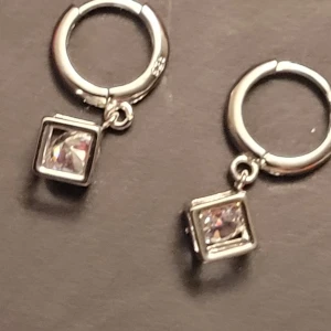 Silvriga örhängen med kubformad sten - Säljer ett par stilrena silvriga örhängen med rund ring och hängande kubformad klar sten. Designen är modern och minimalistisk, perfekt för dig som gillar att sticka ut med detaljer. Stenarna glittrar snyggt i ljuset och ger en cool touch till din look.