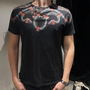 Marcelo Burlon T-shirt  - Svart t-shirt från Marcelo Burlon med ett fett tryck av grå och röda ormar över axlar och bröst. Rund hals och korta ärmar. Materialet är mjuk bomull som sitter skönt mot kroppen. Perfekt för dig som gillar edgy streetwear och vill sticka ut.