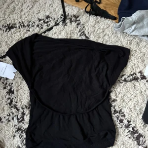 Svart offshoulder topp från Bik Bok - Säljer en svart rygglös topp från Stein  i mjuk bomull. Toppen har en tajt passform och ärmlös design med breda axlar som ger en cool och avslappnad vibe. Perfekt att styla med jeans eller kjol för en trendig look.