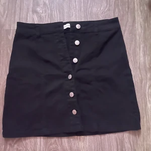 Svart kortkjol från Jacqueline de Yong - Svart kortkjol från Jacqueline de Yong i storlek S. Kjolen har en rak passform och är dekorerad med silvriga knappar framtill. Tillverkad i mjukt bomullstyg som känns skönt mot huden. Perfekt för dig som gillar enkel och stilren design.