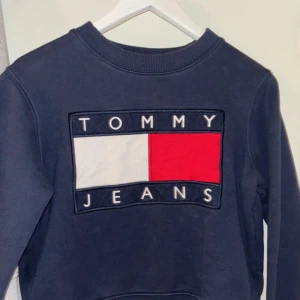 Blå sweatshirt från Tommy Jeans - Snygg mörkblå sweatshirt från Tommy Jeans i storlek S. Tröjan har ett stort broderat logotyptryck i vitt, rött och blått på bröstet. Rund halsringning och ribbade muddar vid ärmslut och nederkant. Perfekt för en chill och stilren look.