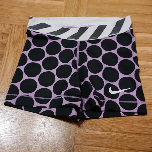 Nike Pro prickiga träningsshorts XS - Säljer ett par Nike Pro träningsshorts i XS med svart och lila prickigt mönster. De har en vit midja med svarta diagonala ränder och är gjorda i ett stretchigt Dri-FIT syntetmaterial som andas. Perfekta för gymmet eller sport.