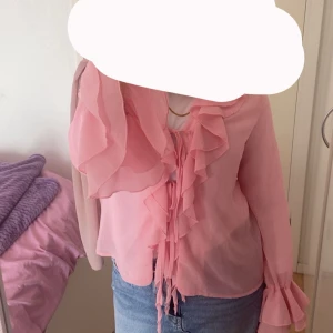 Rosa blus med volanger och snörning - Säljer en superfin rosa blus med långa ärmar, volanger längs halsringningen och framtill, samt snörning vid bröstet. Blusen är gjord i ett lätt och luftigt material som ger en romantisk vibe. Perfekt att styla med jeans eller kjol för en söt look.