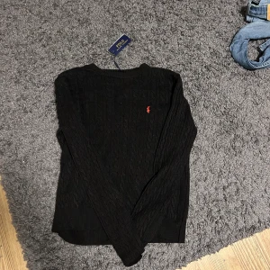 Svart kabelstickad tröja Polo Ralph Lauren - Svart kabelstickad tröja från Polo Ralph Lauren i storlek S. Klassisk design med rund hals och långärmad passform. Tröjan har det ikoniska röda Polo-logot broderat på bröstet och är tillverkad i mjuk bomull som känns skön mot huden. Hör av er vid funderingar 