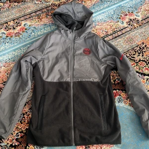 Nike PSG fleecejacka med huva, grå/svart - Nike PSG fleecejacka i grått och svart med huva och hel dragkedja. Jackan har PSG-logga på bröstet och Nike Swoosh på ärmen. Tillverkad i mjuk fleece och polyester, perfekt för kyliga dagar. Två sidofickor och sportig look.
