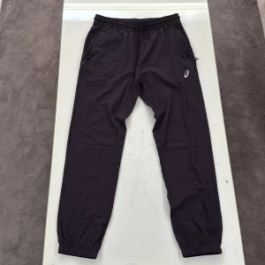 Svarta byxor Asics - Svarta Asics träningsbyxor med elastisk midja och snörning. Byxorna har smal passform, resår vid bensluten och praktiska fickor med dragkedja. Materialet är lätt och syntetiskt, perfekt för sport och aktivitet. 