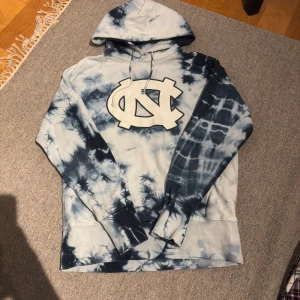 Blå tie-dye hoodie UNC - Säljer en blå och vit hoodie från champion  med stor logga framtill. Tröjan har bara använts 2-3 gånger. Tröjan har huva med snörning och långa ärmar.                                   Nypris: 2000kr