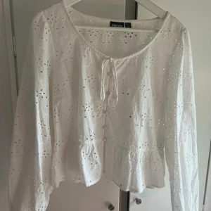 Vit blus med broderade detaljer från Pieces - Säljer en vit blus från Pieces i storlek L. Blusen har broderade hålmönster, volang nedtill och knytband vid halsen. Långärmad och tillverkad i mjuk bomull, perfekt för vår och sommar. Söt och luftig modell som ger en fräsch vibe.