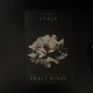 Crave - En mörk och lockande bok med en dramatisk omslagsbild av en vit blomma med blodstänk. Perfekt för dig som gillar mystik, spänning och romantik med en twist. Skriven av New York Times-bästsäljande författaren Tracy Wolff.