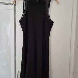 Klänning - Svart ärmlös klänning från H&M i storlek M. Klänningen har mesh-detaljer vid axlarna och en klassisk rund halsringning. Modellen är kort och har en lätt utställd kjol som ger en snygg siluett. Perfekt för dig som gillar stilrena och enkla plagg.