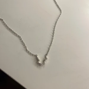 Silverhalsband med stjärnformade stenar - Ett stilrent halsband i silverfärgad metall med en tunn kedja och ett hänge format som en liten båge med fem gnistrande stjärnformade stenar. Perfekt för dig som gillar diskreta men ändå glittriga detaljer.