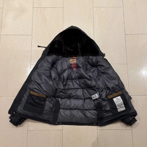 Svart dunjacka Parajumpers Special Edition XS - Säljer en svart Parajumpers Special Edition dunjacka med huva och skinnärmar. Jackan har flera fickor, patch på axeln och coola detaljer som knappar och dragkedjor. Insidan är quiltad och fodrad med mjukt material. Perfekt för dig som vill ha en unik och varm jacka.