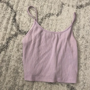Ljuslila ribbad linnetopp - Säljer en ljuslila ribbad linnetopp med smala axelband. Toppen har en croppad passform och är perfekt för varma dagar. Materialet är mjukt och stretchigt, vilket gör den skön att bära. Enkel och stilren design som är lätt att matcha.