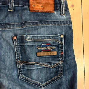 Tommy Hilfiger blå jeans - Äkta Tommy Hilfiger jeans i mörk denim med vintage/slitna detaljer. Snygg broderad logga bak. Bra skick med lite slitage längst ner (se bild).