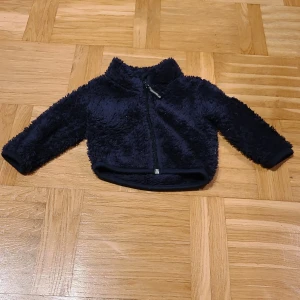Mörkblå teddyjacka från Little Star - Mysig mörkblå teddyjacka från Little Star i storlek 68. Jackan har fluffig yta, dragkedja framtill och långärmade ärmar. Perfekt för kyliga dagar och har en liten reflexdetalj på ryggen. Snygg och enkel att matcha med andra plagg.