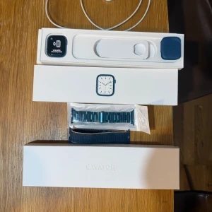 Apple Watch Series 7 blå aluminium - Apple Watch Series 7 med blå aluminiumboett och två olika blå armband, ett i metall och ett i textil. Klockan har en fyrkantig form och levereras med originalförpackning och laddare. Perfekt för dig som gillar smarta funktioner och stilren design.