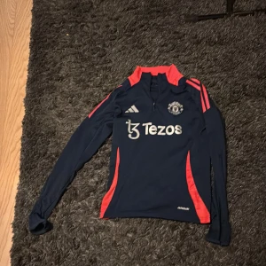 Manchester United långärmad träningströja - Snygg långärmad Manchester United träningströja från Adidas i mörkblått med röda detaljer och halv dragkedja. Tröjan har klubbmärke och Adidas-logga på bröstet samt Tezos sponsortryck. Tillverkad i lätt och funktionellt material.