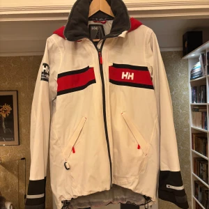 Helly Hansen vit seglarjacka (ish Medelstorlek)  - Snygg seglarjacka från Helly Hansen i vitt med röda detaljer. Jackan har hög krage med fleecefoder, dragkedja framtill, två fickor med dragkedja och justerbar nederkant. Perfekt för blåsiga dagar på stan eller en tur på sjön. Jackan är ganska liten för sin storlek. Hade passat bättre för S eller M personer. 