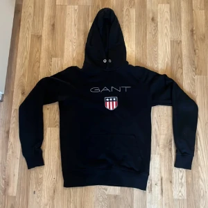 Svart hoodie från GANT med logga - Svart hoodie från GANT i storlek S med stor broderad logga och emblem på bröstet. Tröjan har huva med snörning, magficka och långa ärmar. Materialet är mjuk bomull och polyester, perfekt för chill dagar. Klassisk och stilren design med vita och röda detaljer.