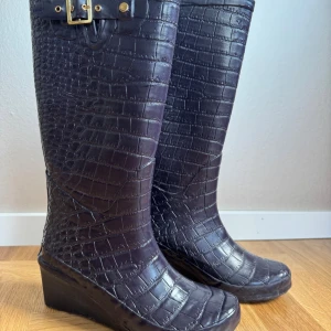 Khombu mörklila krokopräglade gummistövlar - Var snygg i slasket! Mörklila, nästan svarta stövlar från Khombu med krokodilpräglad yta och guldfärgat spänne upptill. Insidan har ett leopardmönstrat foder som ger extra edge. Stövlarna har kilklack som gör dem sköna att gå i och är tillverkade i syntetmaterial. 