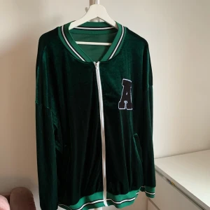 Grön bomberjacka med A-patch - Snygg mörkgrön bomberjacka i mjuk sammet med vita och gröna ränder på krage och muddar. Jackan har en vit dragkedja framtill och en stor A-patch på bröstet. Perfekt för dig som gillar sportig och chill stil.