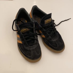 Adidas Handball Spezial svart/orange 38⅔ - Säljer ett par Adidas Handball Spezial sneakers i svart mocka med orangea/brandgula detaljer och klassiska tre ränder på sidan. Skorna har gummisula, snörning och guldfärgad Spezial-logga. Insidan är dekorerad med Adidas-tryck..
