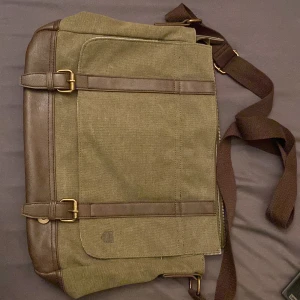 Brun datorväska från Pier One - Snygg datorväska från Pier One i brunt tyg och skinn med detaljer i mässing. Väskan har justerbar axelrem, praktiska fack och randigt innerfoder. Perfekt för att bära laptop och böcker, med stilren design och smarta spännen framtill.