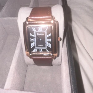 Geduo fyrkantig klocka brun/svart - Stilren fyrkantig klocka från Geduo med svart urtavla, romerska siffror och brunt läderband. Boetten är i guldtonad metall och klockan har klassisk quartz-mekanism. Perfekt accessoar för dig som gillar en tidlös look. Pris kan diskuteras 